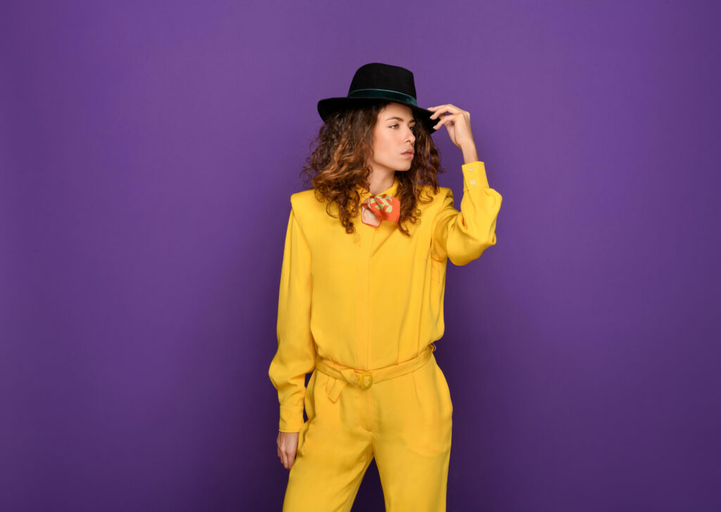 Fashionable woman in bright yellow suit and black hat against a purple background.