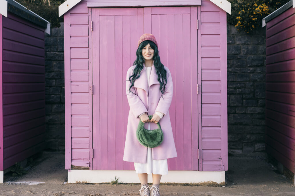 Woman dressed in all pink vintage clothing in front of a pink beach hut.